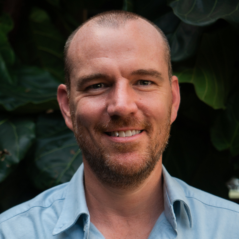 Man with shaved head and beard smiling outdoors against dark leafy background.